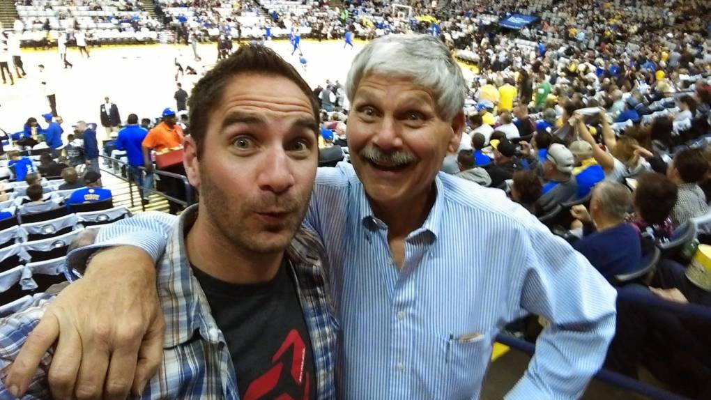 a young man in a flannel shirt and an older man, his father, smile for the camera with their arms around each other at a basketball game
