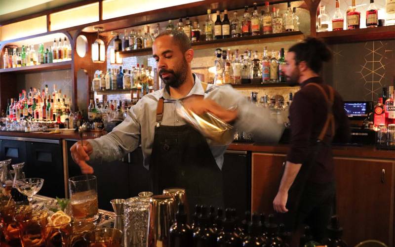 A bartender in an apron vigorously shakes a a tumbler to make a cocktail. A fully stocked bar is visible behind him.