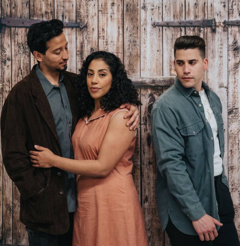 a photo of three people posed in front of a wooden door, two youngish Latino-appearing men with a Latina woman in an orange standing between them, looking at the camera