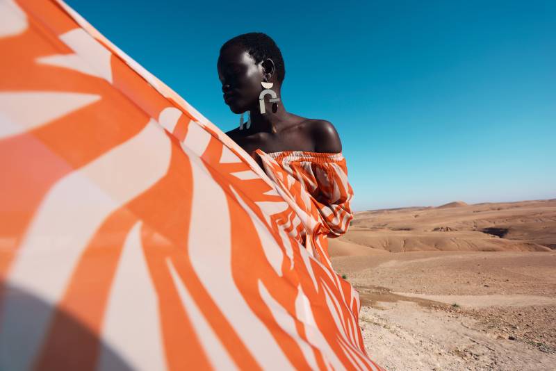 a tall Black model wears a white and orange patterned dress against a blue sky