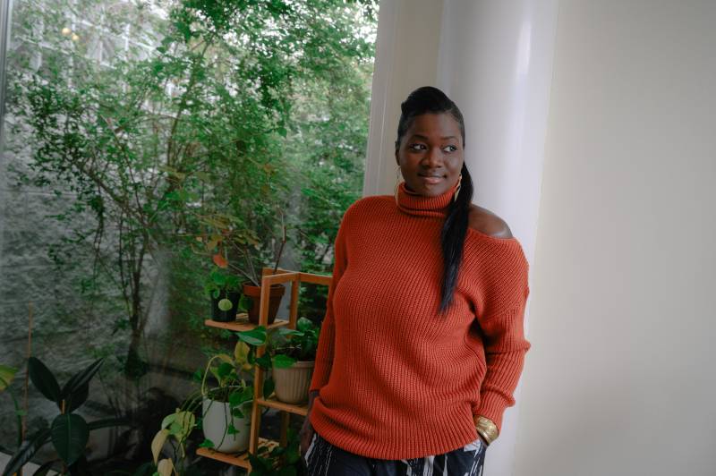 a young Black woman in an orange sweater stands smiling in a house filled with plants