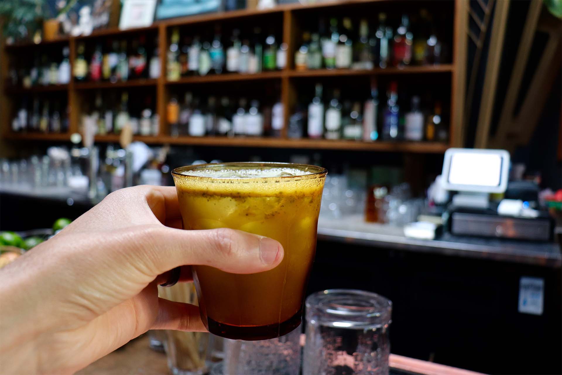 A hand holding up a cocktail, with fruity, pulpy bits floating on top. A fully stocked bar is visible in the background.
