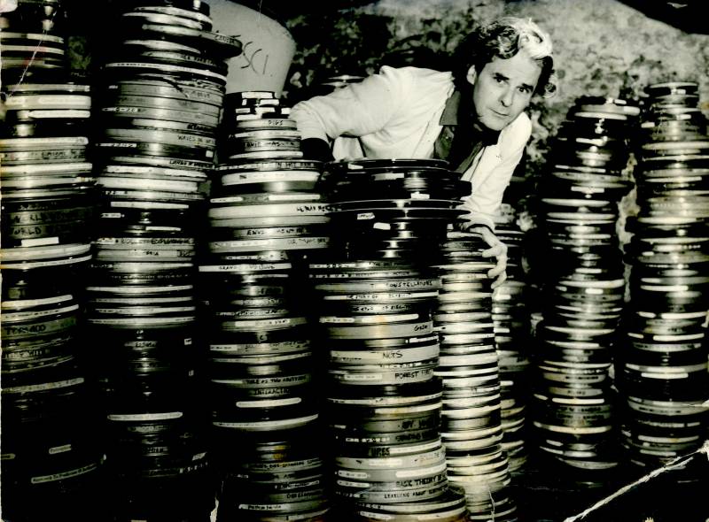 a black and white photo of a man in a white suit jacket peering out from behind a stack of film reels