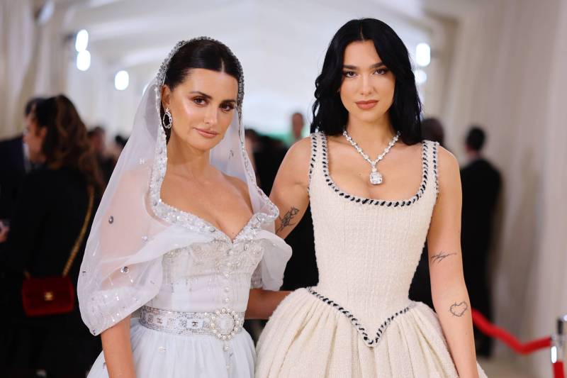 Two women with pale flawless skin and dark brown hair stand side by side. One is wearing a corseted white dress with black stitching. The other is a wearing a plunging, belted white dress with a transparent white hood.