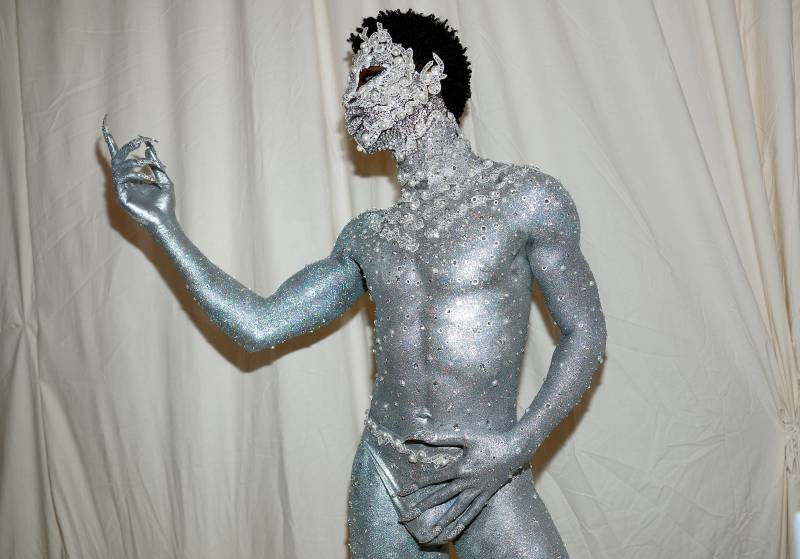 A man in silver body paint and crystal-encrusted face mask strikes a pose in front of a white curtain. He is wearing only a loin cloth.