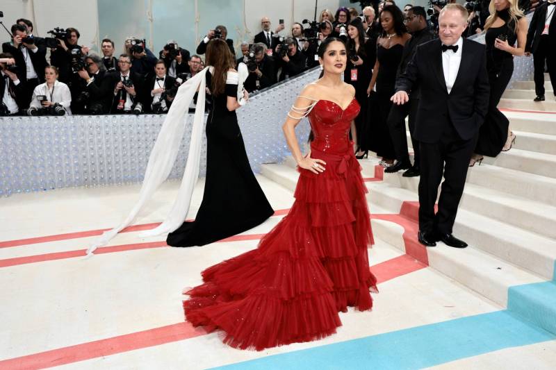 A striking movie star stands on a red carpet wearing a red, tiered gown and bodice, hand on her hip. Photographers are lined up behind her.