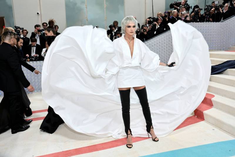 A white woman with cropped shaggy silver hair stands on a red carpet surrounded by photographers, fanning out a huge white cape from a mini dress. Her legs are covered with black leather sheaths.