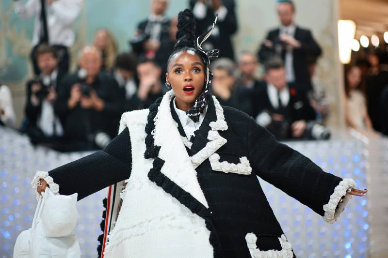 A Black woman wearing a stunned facial expression stands wearing an oversized conical black and white suit coat, held up by a wired underskirt.