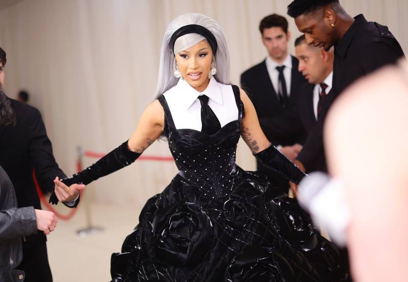 A Black woman wearing a dramatic black gown with white sleeveless collared shirt and black tie underneath, stands on the red carpet, gloved arms outstretched. She is wearing a long, straight, silver wig.