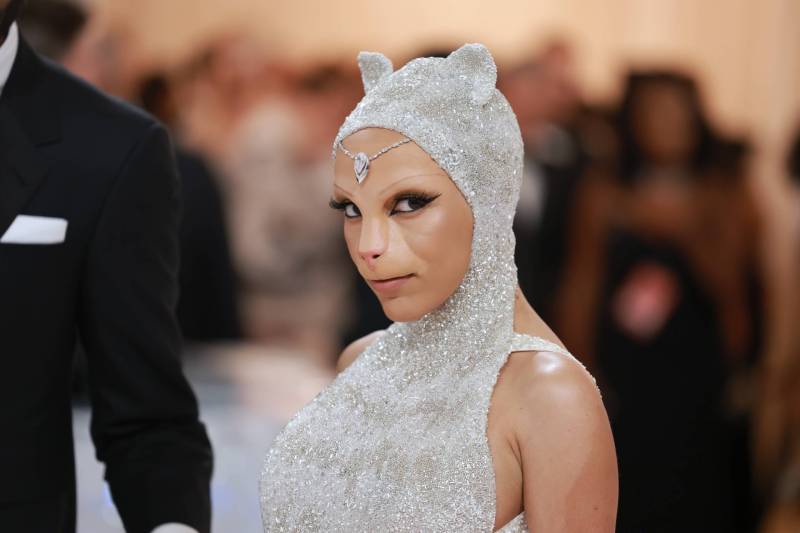 A Black woman wearing face enhancements that make her look like a cat poses on the red carpet. She's wearing a silver gown with hood that has cat ear embellishments.