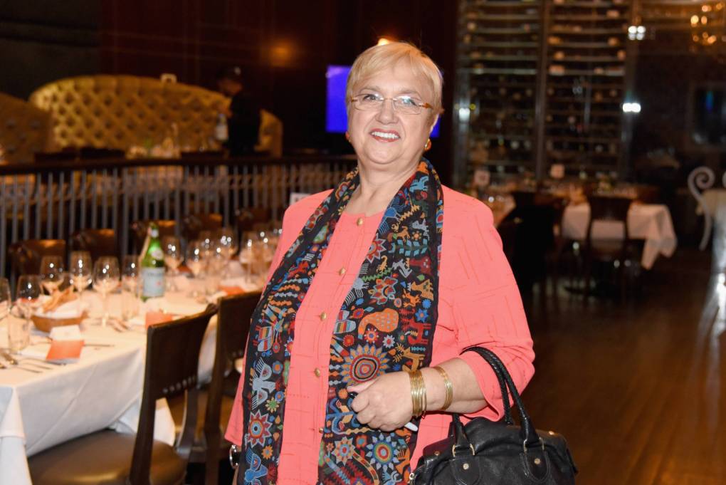 A woman in her sixties stands in an empty restaurant, smiling broadly, handbag resting on forearm.