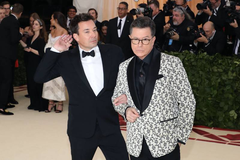 Two men in tuxedos pose self-consciously and ironically on a red carpet, rows of photographers lined up behind them.