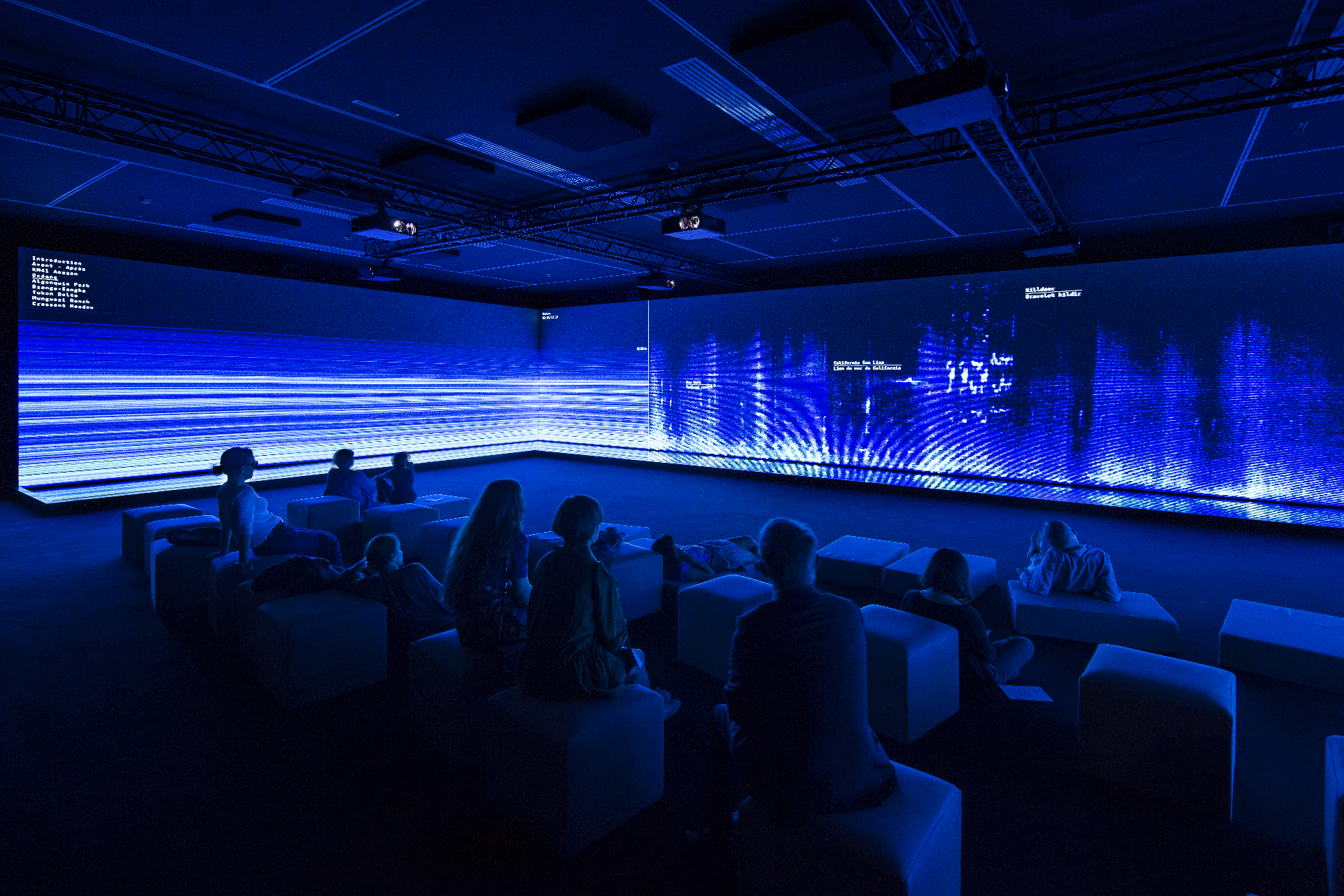 Dark room with seated audience looking at wide corner of blue data-like lines on video screen