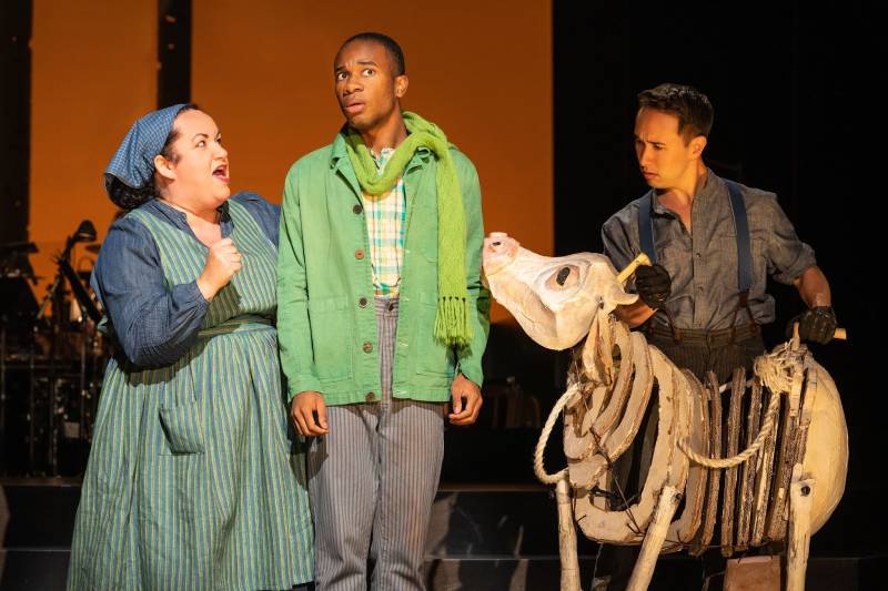 three people, a white woman, a Black man and a Japanese-American man in colorful outfits, perform on a stage with a fake animal skeleton of some kind