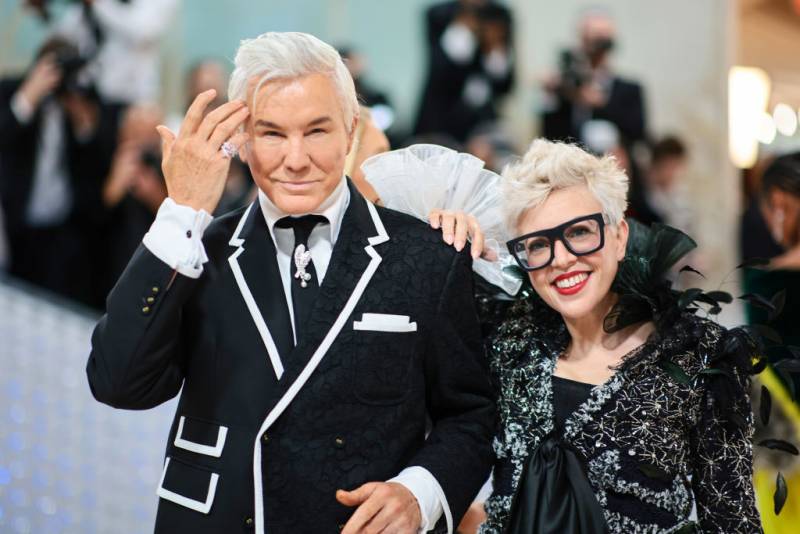 A man with white hair and a black-and-white suit with a woman in black wardrobe and glasses.