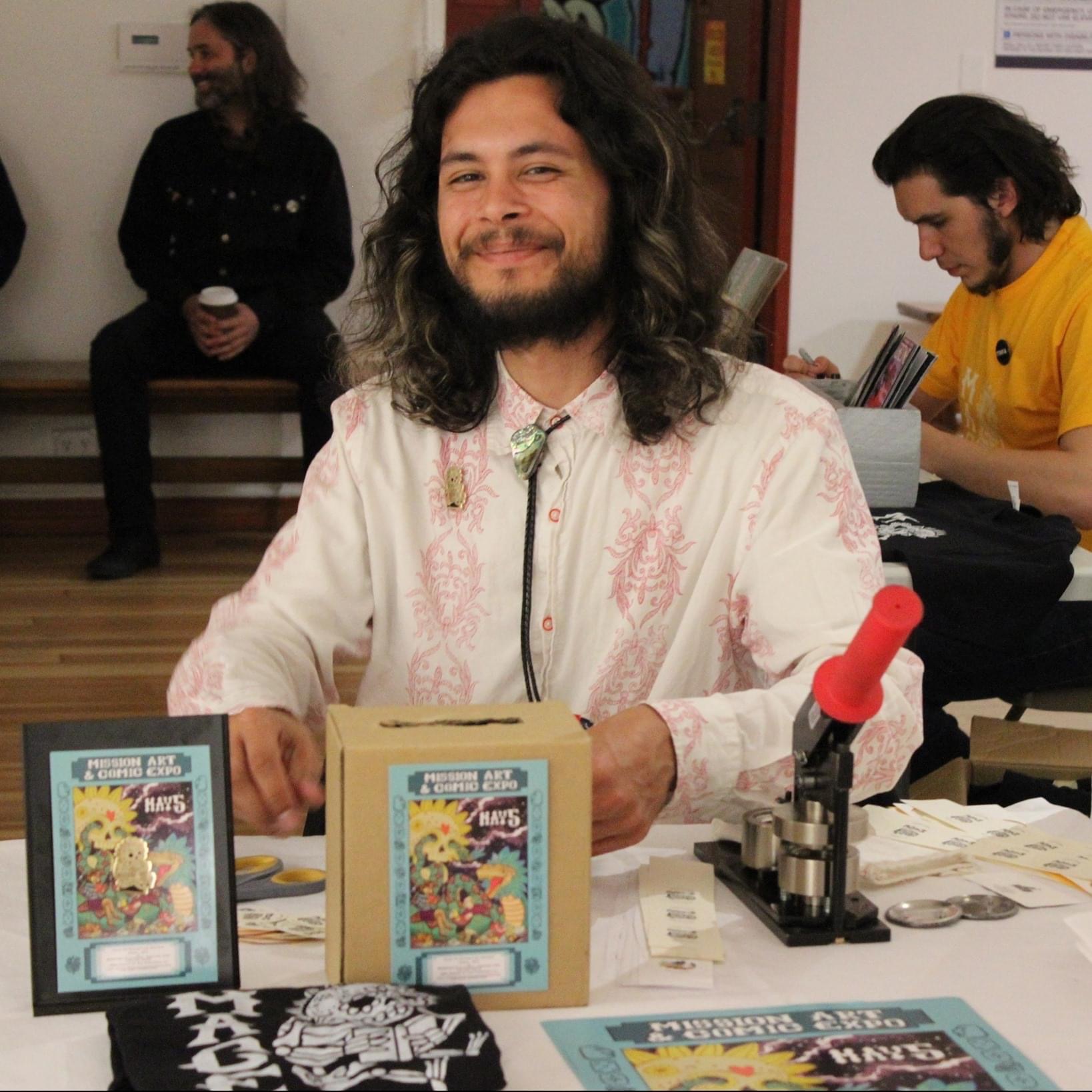 Smiling person with long dark hair, bolo tie in front of table of zines and buttons, two people in background