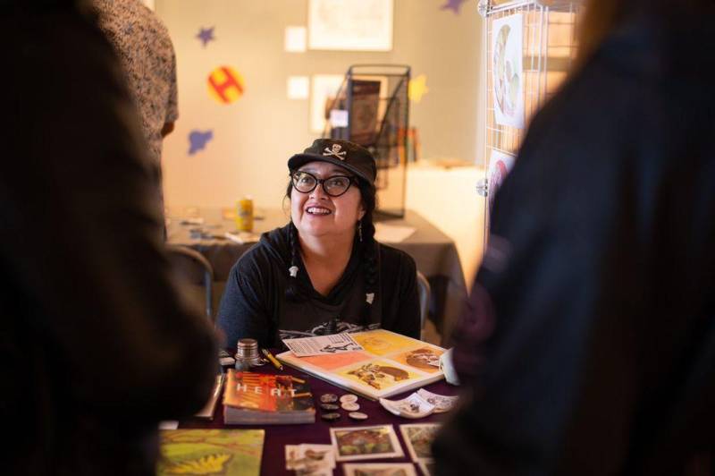 Woman in black ballcap and black rimmed glasses looks up from table of illustrations and printed materials