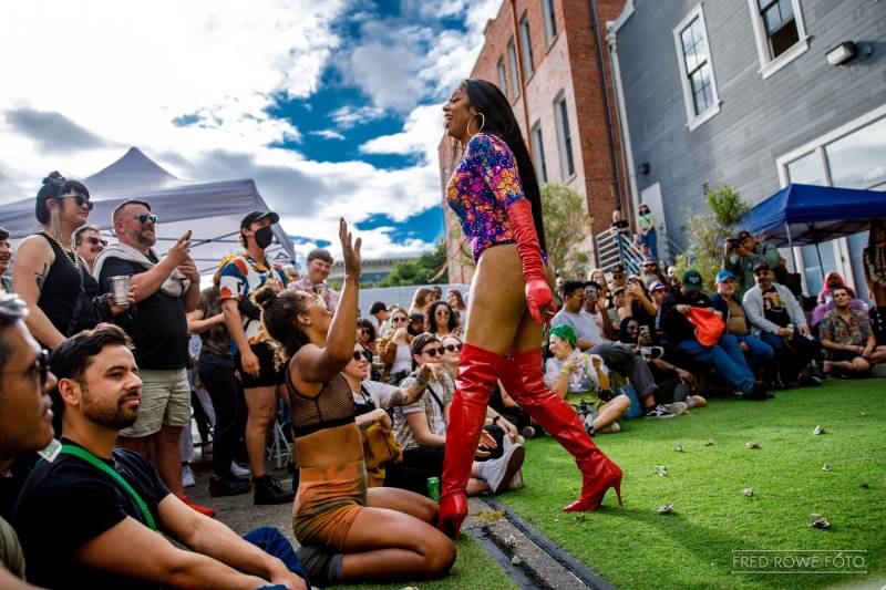 a drag performer in high heel red boots laughs as she performs outside for a crowd