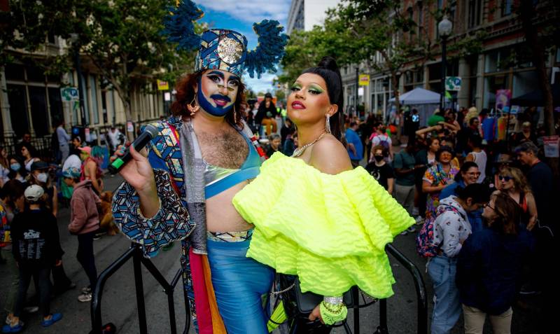 two colorfully dressed drag performers pose for the camera while people fill the street behind them at a drag festival