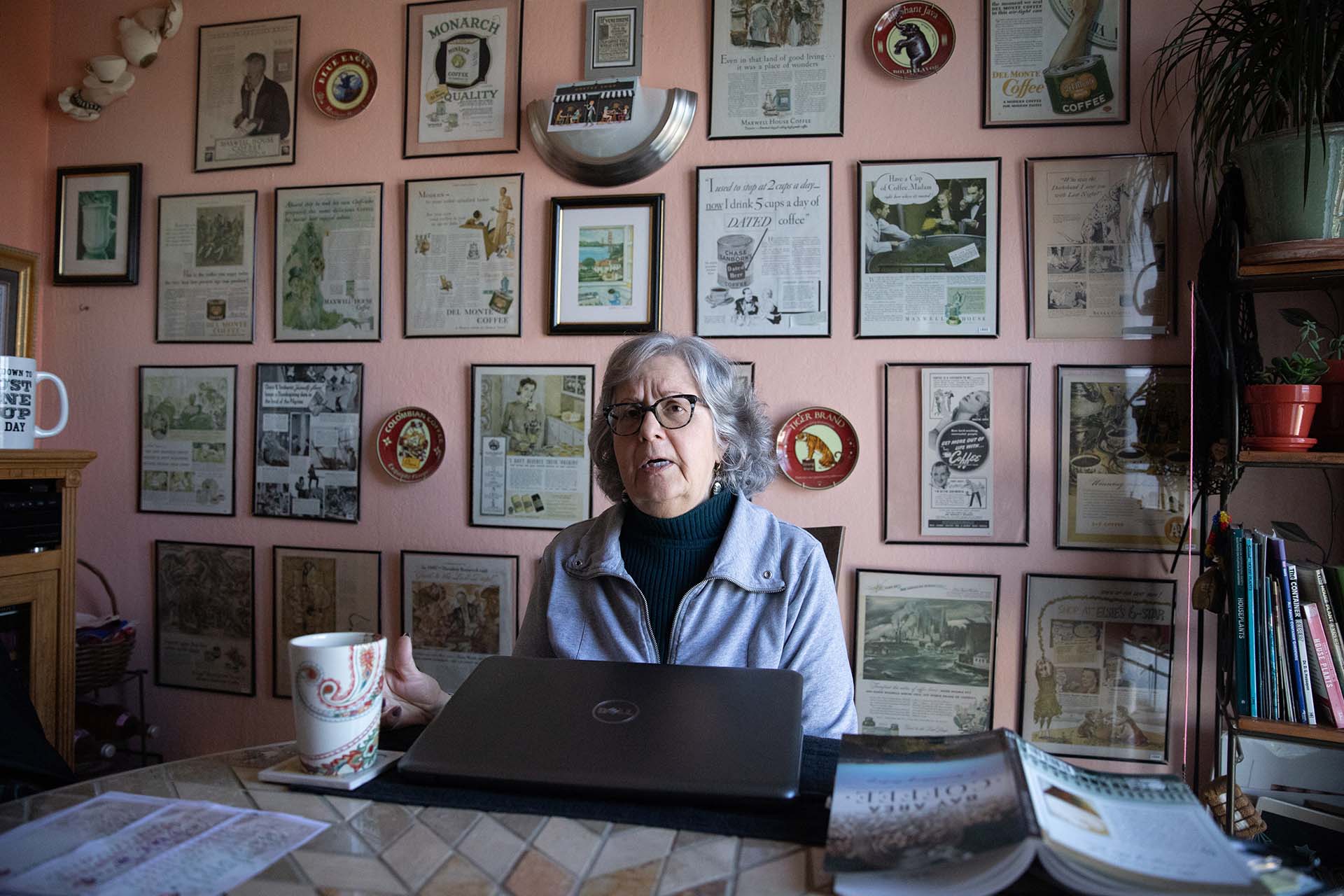 Woman wearing glasses sits in front of a wall covered in old newspaper clippings and coffee advertisements.