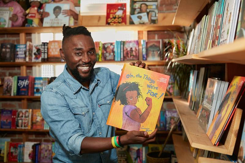 a young Black man in a denim button up shirt smiles while holding a children's book in a bookstore