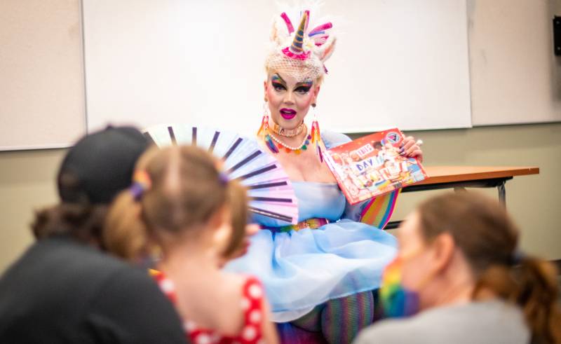 a drag performer in a blonde wig and colorful dress reads to kids in a classroom