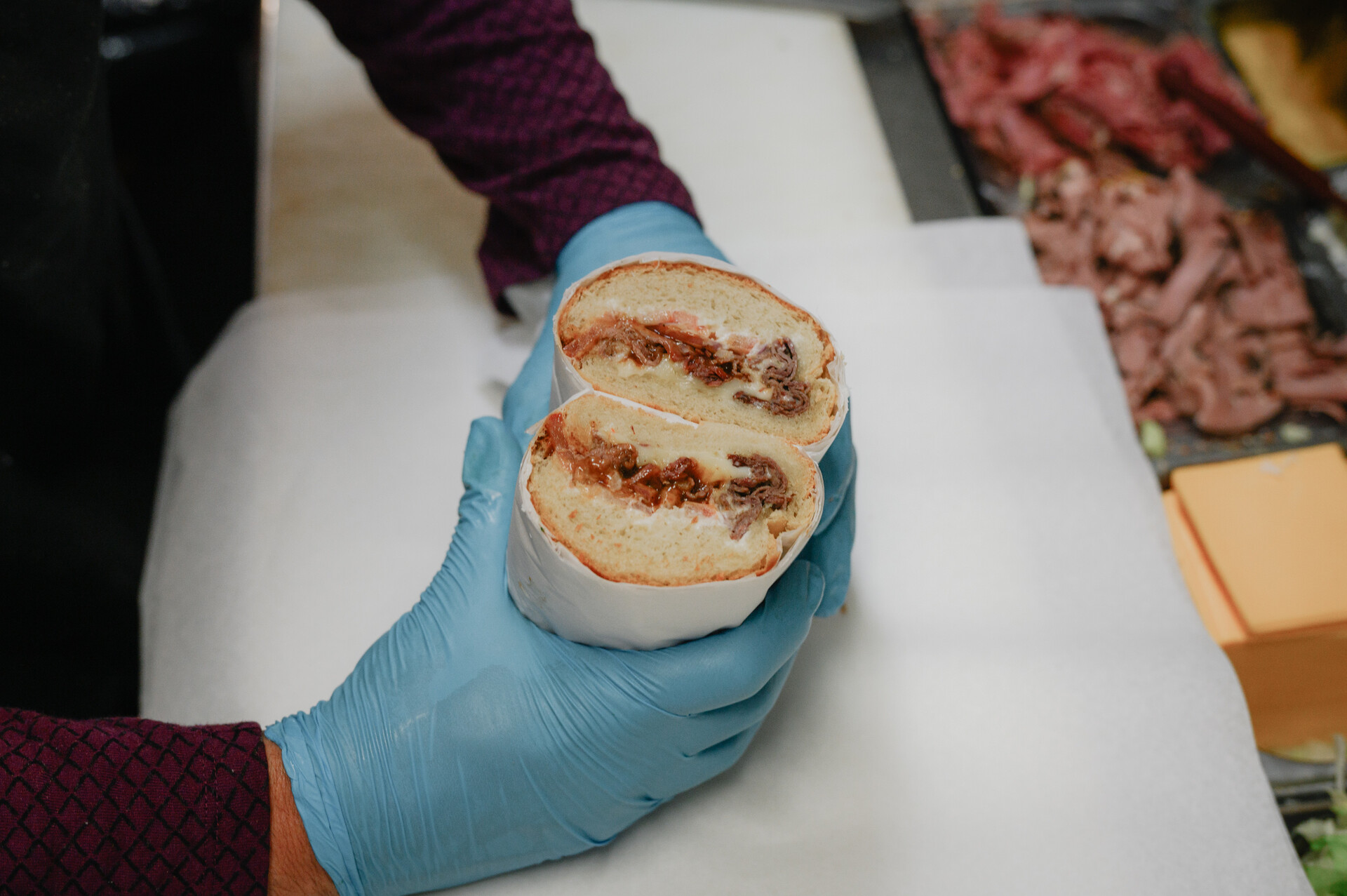 A deli worker wearing blue kitchen gloves holds a pastrami sandwich, cut so that the meaty cross section is visible.