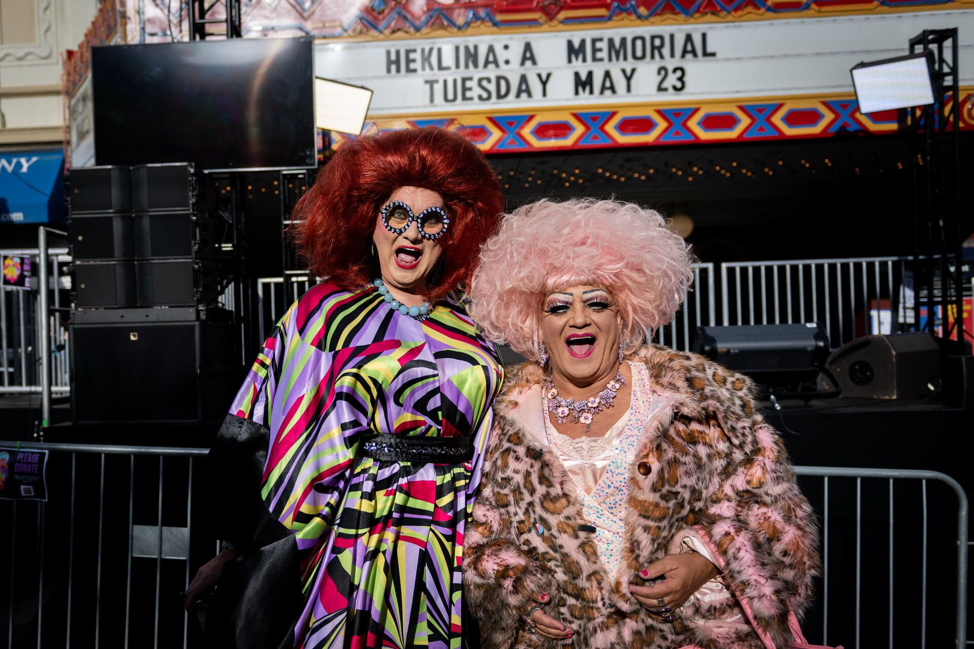 Two drag performers, dressed up in very colorful wigs and puffy dresses, smile at the camera.