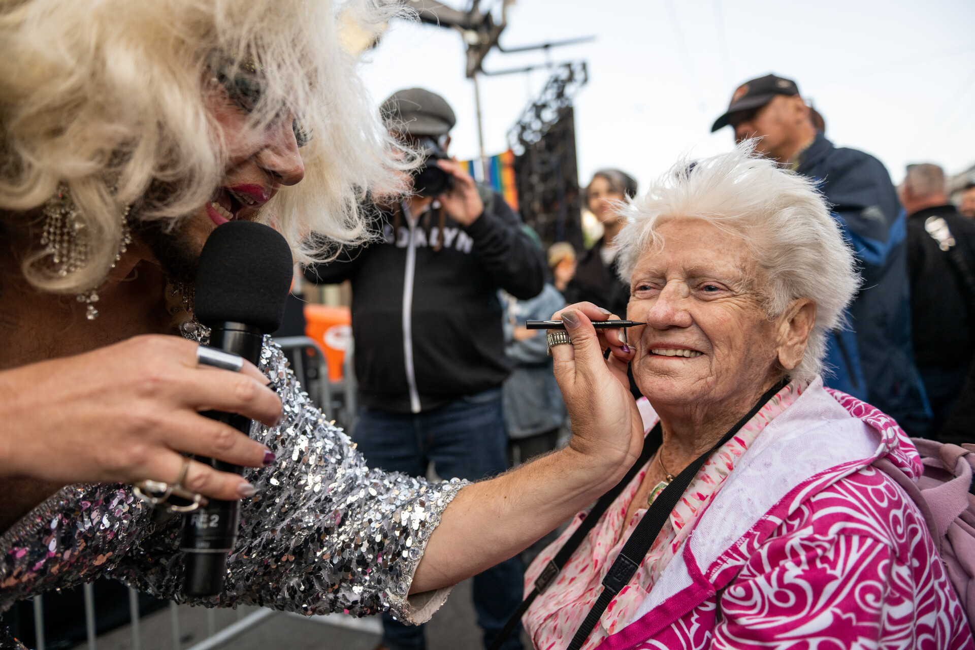 A drag performer holds a microphone in one hand, and with the other, holds a marker and draws a beauty mark on the face of a woman standing in the crowd outside the Castro Theater.