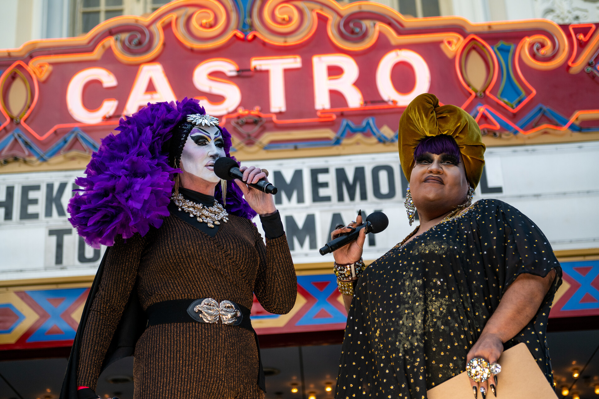 Two drag artists speak with microphones to a large crowd in front of San Francisco's Castro Theater.