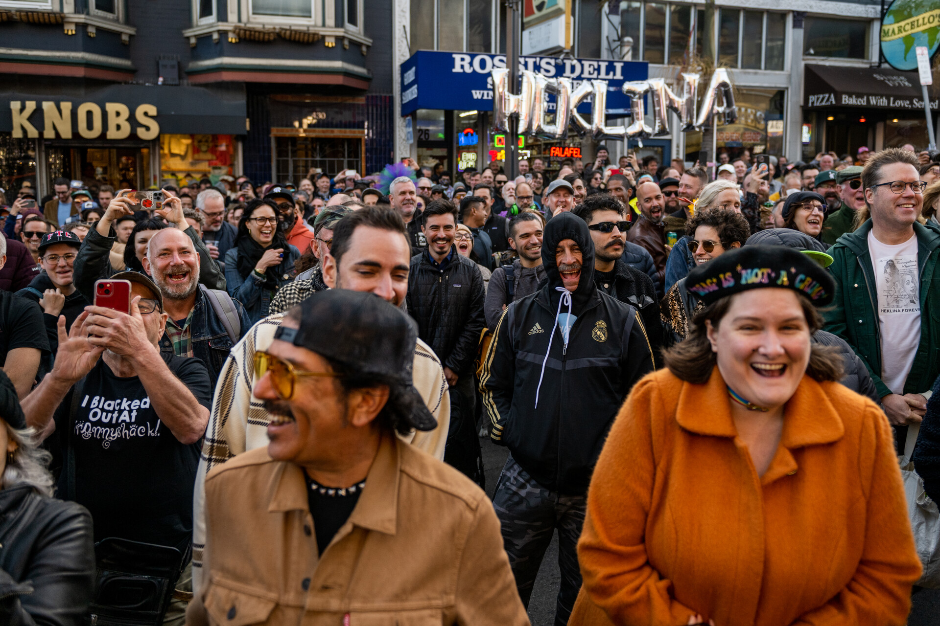 A crowd of hundreds, of all ages, stands outside on San Francisco's Castro Street and laughs, many with smiles on their faces. In the background, there are balloons in the shape of the letters that spell out "Heklina."