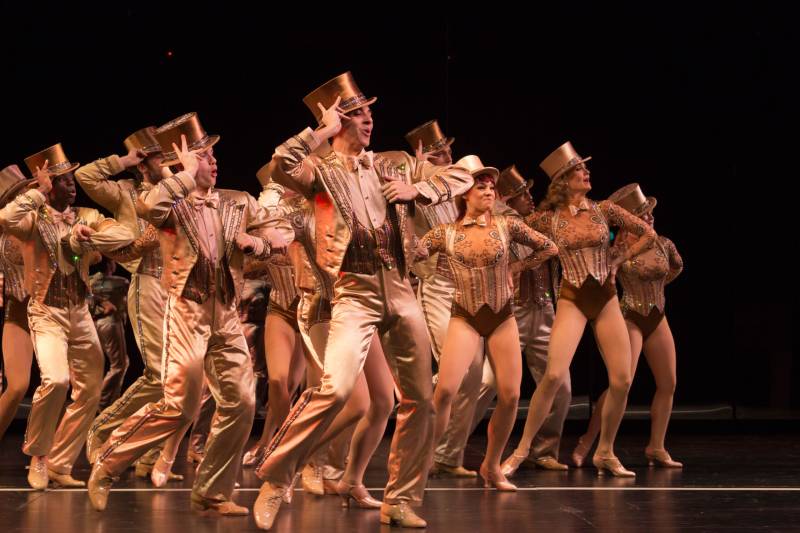 a large group of people in top hats and shiny outfits perform a dance in a chorus line on stage