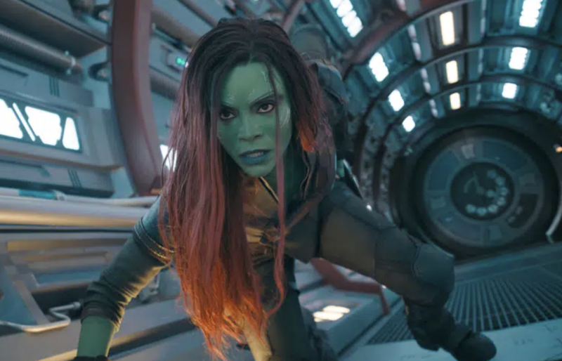 A woman with long auburn hair and a green face adopts a fight position close to the ground within a space craft.