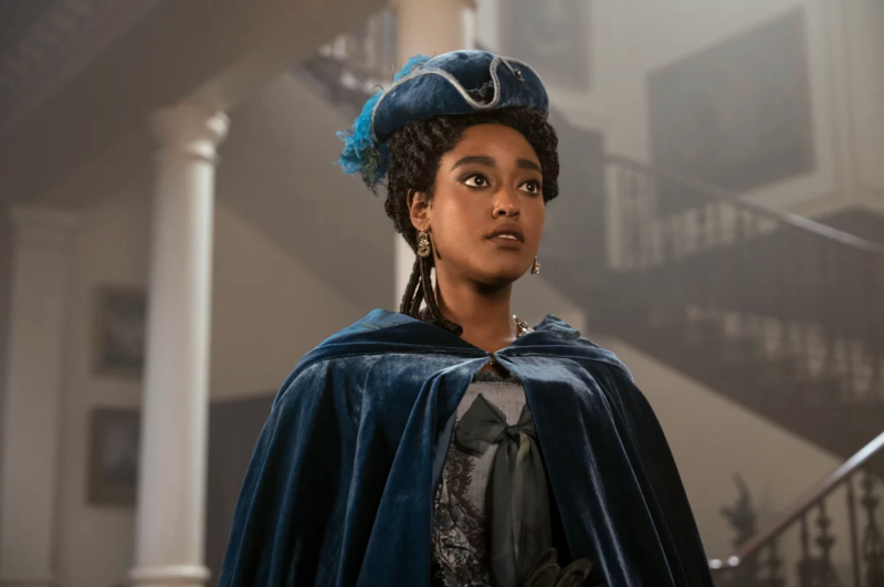 A beautiful young Black woman stands in an ornate hall wearing blue velvet cape and matching hat.