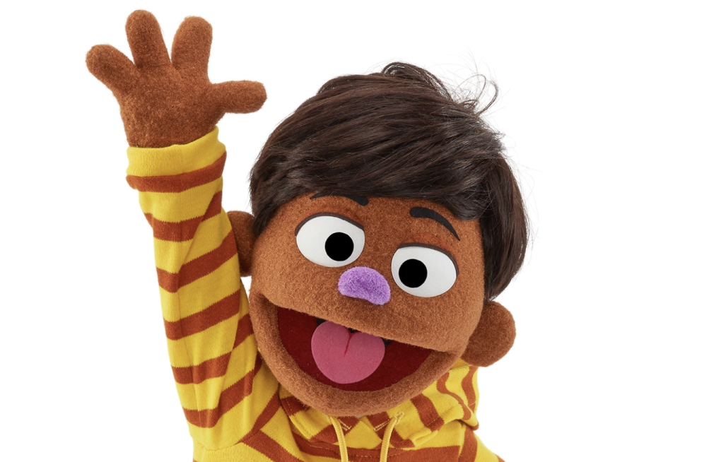 A brown muppet with brown hair and purple nose smiles and waves. He is wearing a yellow and brown hoodie.