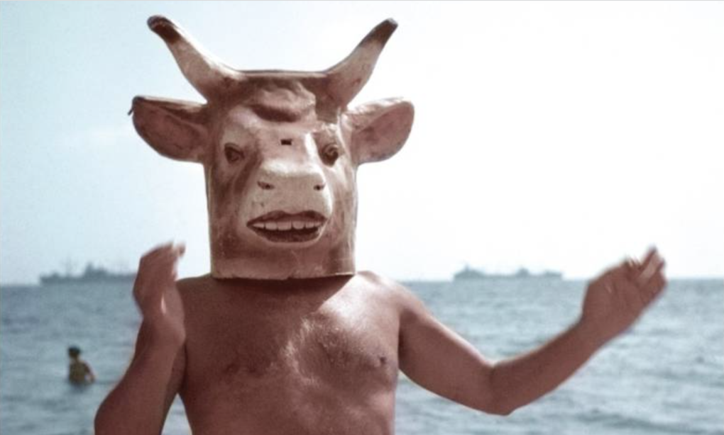 A shirtless man standing on a beach wearing a horned mask.