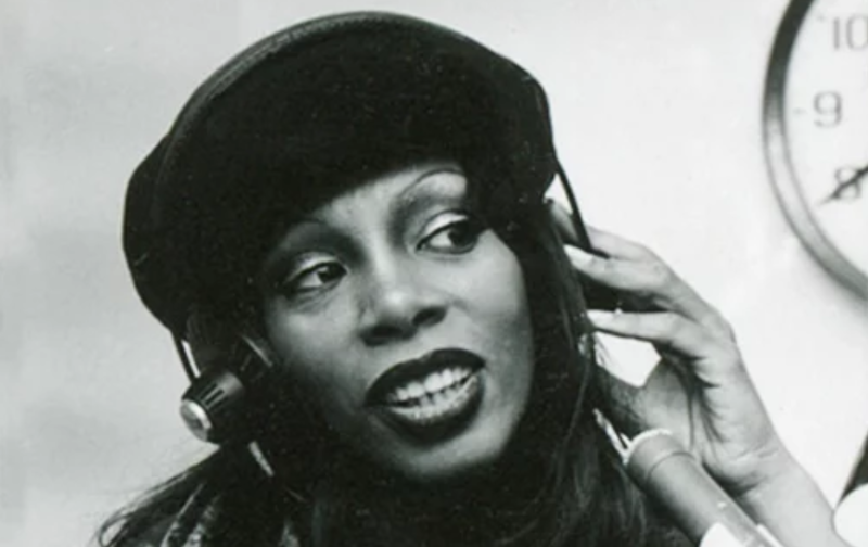 An attractive Black woman wearing a leather jacket and beret looks off to one side, distracted. She is wearing headphones and sitting behind a microphone.