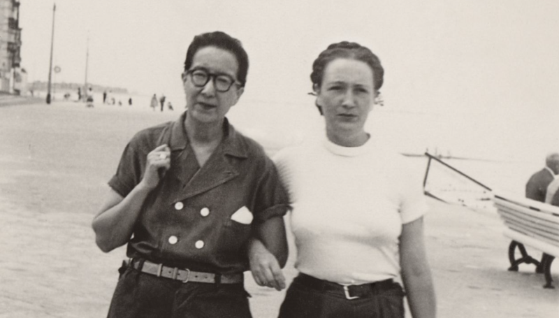 Two women walk arm in arm. One is Asian, wearing round spectacles with short-slicked back hair. The other is white, hair braided neatly around her head, wearing a tight white t-shirt and trousers.