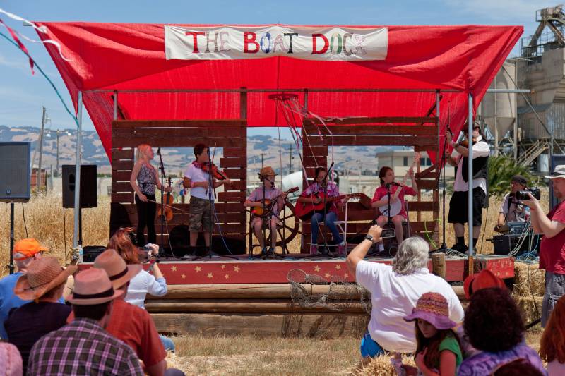 a band made up of five kids with instruments performs on a stage with a sign that reads 'the boat dock' at a festival