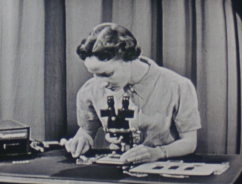 a still from a black and white movie of a woman sitting looking into a microsope