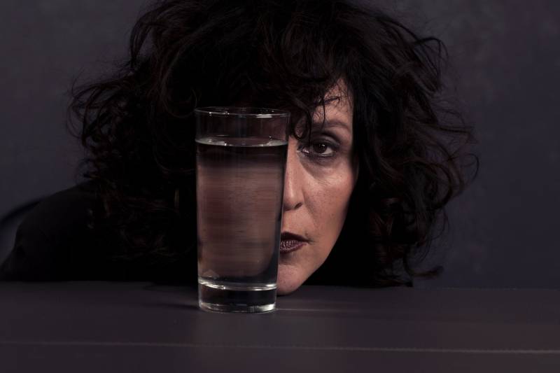 a glass of water on a table in front of a woman's face, lit darkly; she has brown skin and black hair and is wearing dark lipstick