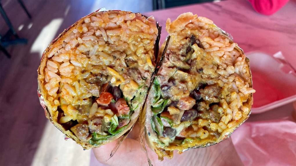 Two halves of a cut-open burrito held so that the cross section is visible. It's stuff full of rice, beans, steak, lettuce, tomato and a creamy golden sauce.