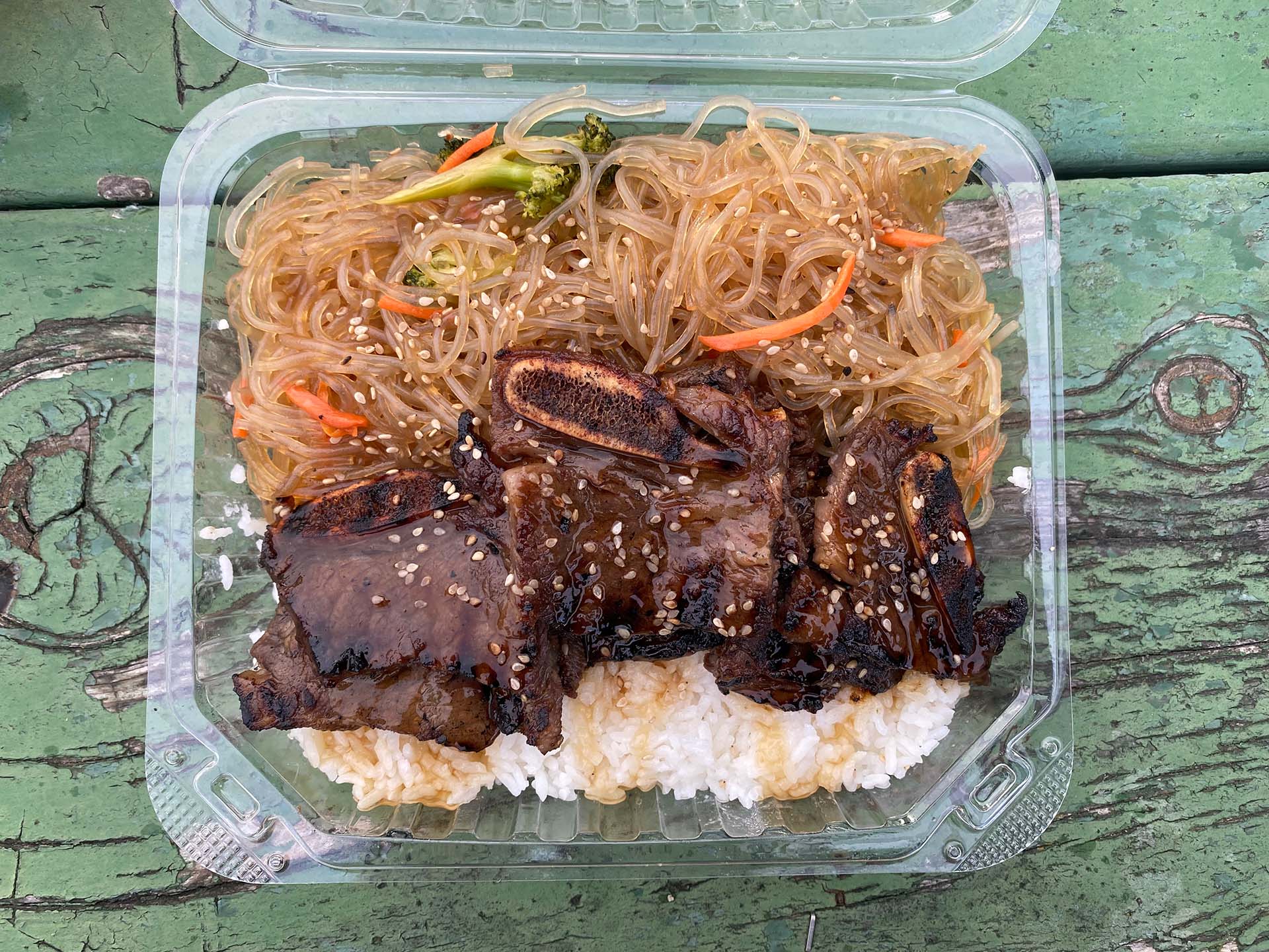 Korean barbecue beef short rib served over rice and japchae (noodles) in a plastic takeout container.