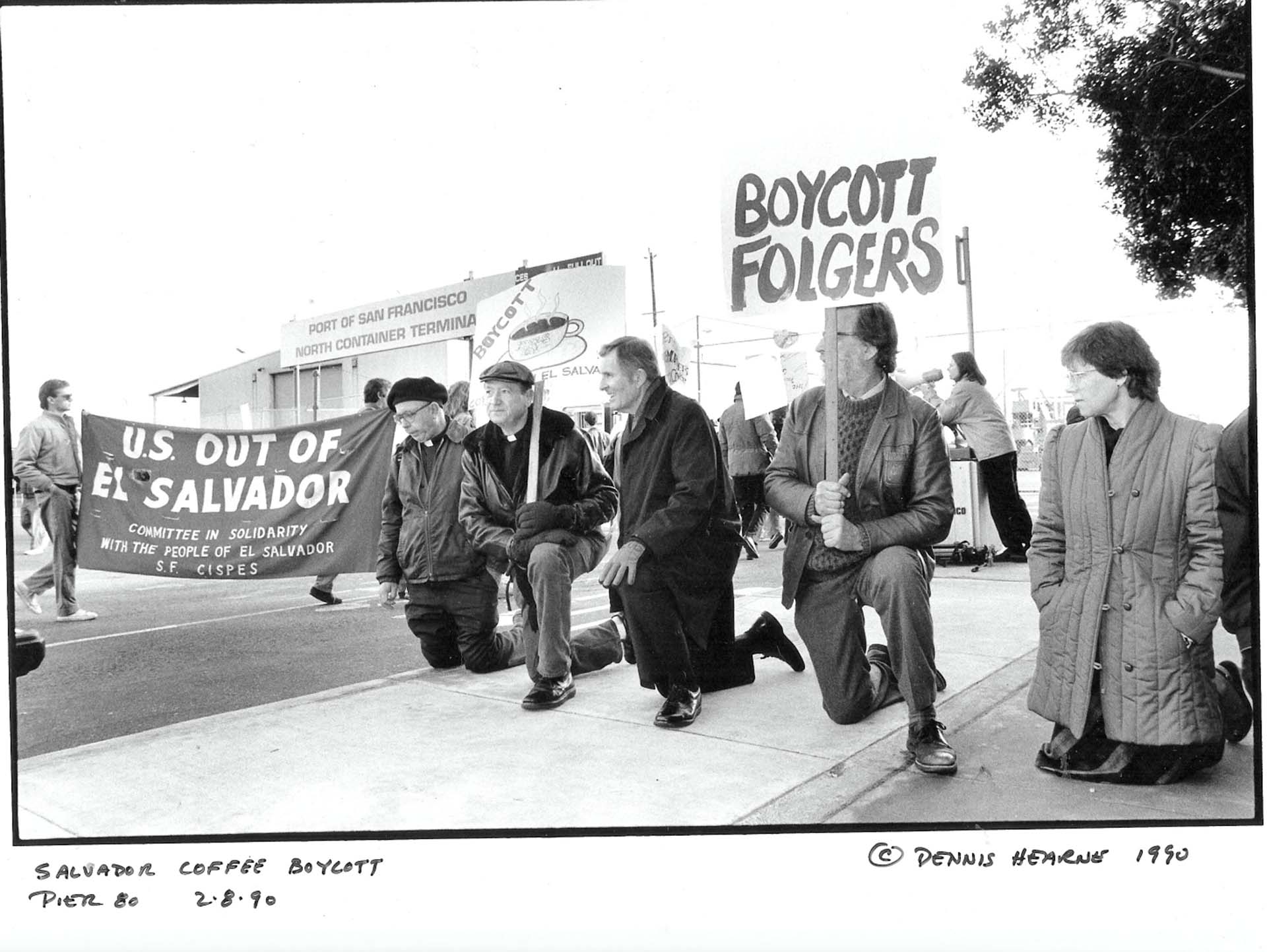 Black-and-white image of protestors taking a knee, holding signs that read, "Boycott Folgers" and "U.S. Out of El Salvador."