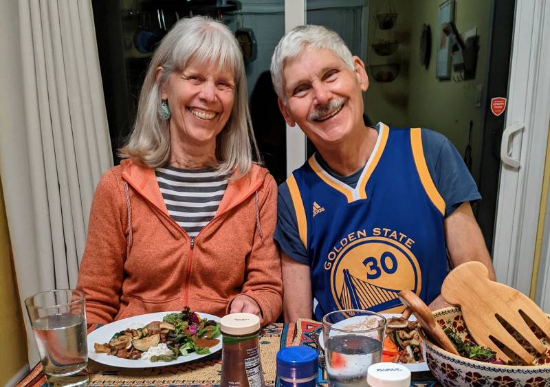 an older woman and man with grey hair smiling at a dinner table. she wears an orange hoodie over a striped black and white shirt and he wears a blue and yellow warriors jersey