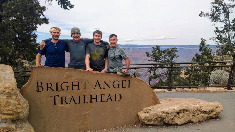 a group of four men, two younger, two older, pose behind a sign that reads 'Bright angel trailhead' 