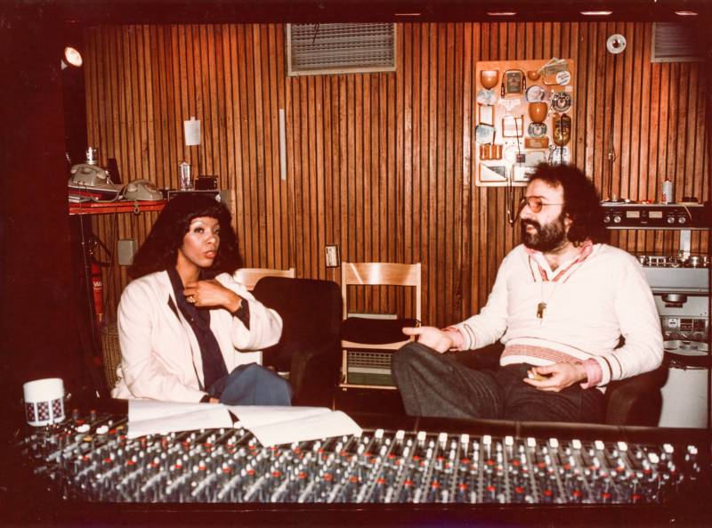 A Black woman and white bearded man sit behind the mixing desk of a recording studio. The walls are wood paneled. 