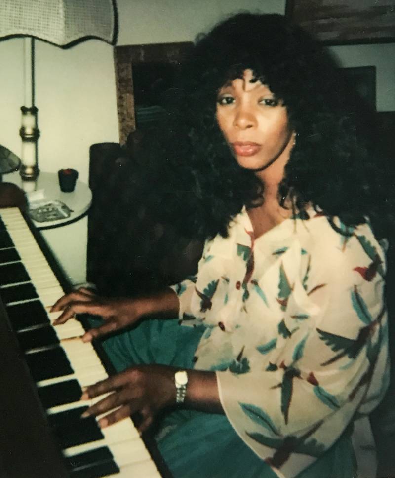 A Black womam sits at a piano, hands on keys, looking sullen.