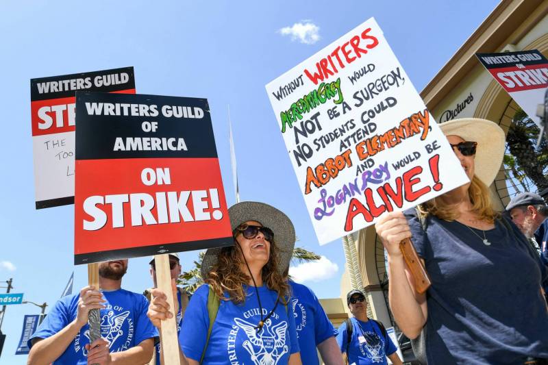 Protesters on a picket line. Signs read: "Writers Guild of America on Strike!" and "Without writers, Meredith Grey would not be a surgeon, no students could attend Abbot Elementary and Logan Roy would be alive!"
