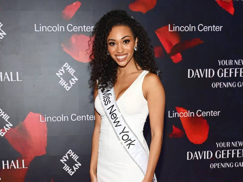 A beautiful young woman of color stands on the red carpet of the Lincoln Center, wearing a white dress and a sash.
